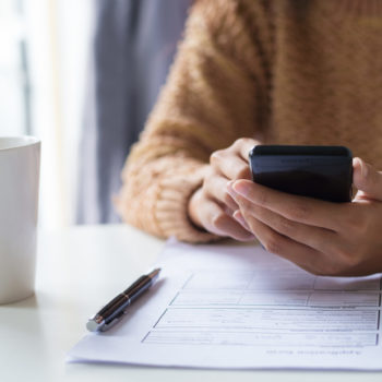 Close-up of business lady using gadget while examining document. Unrecognizable woman in warm sweater sitting at table and checking message. Technology concept
